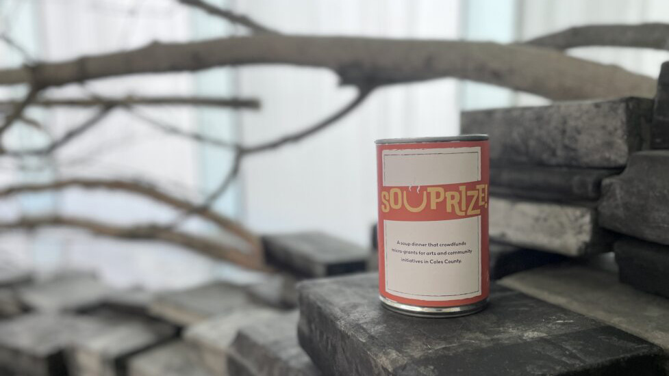 A red soup can labelled "Souprize!" is set on top of a blackened, burned book stack in Marie Bannerot Mcinerney's "Falling Into Milk"
