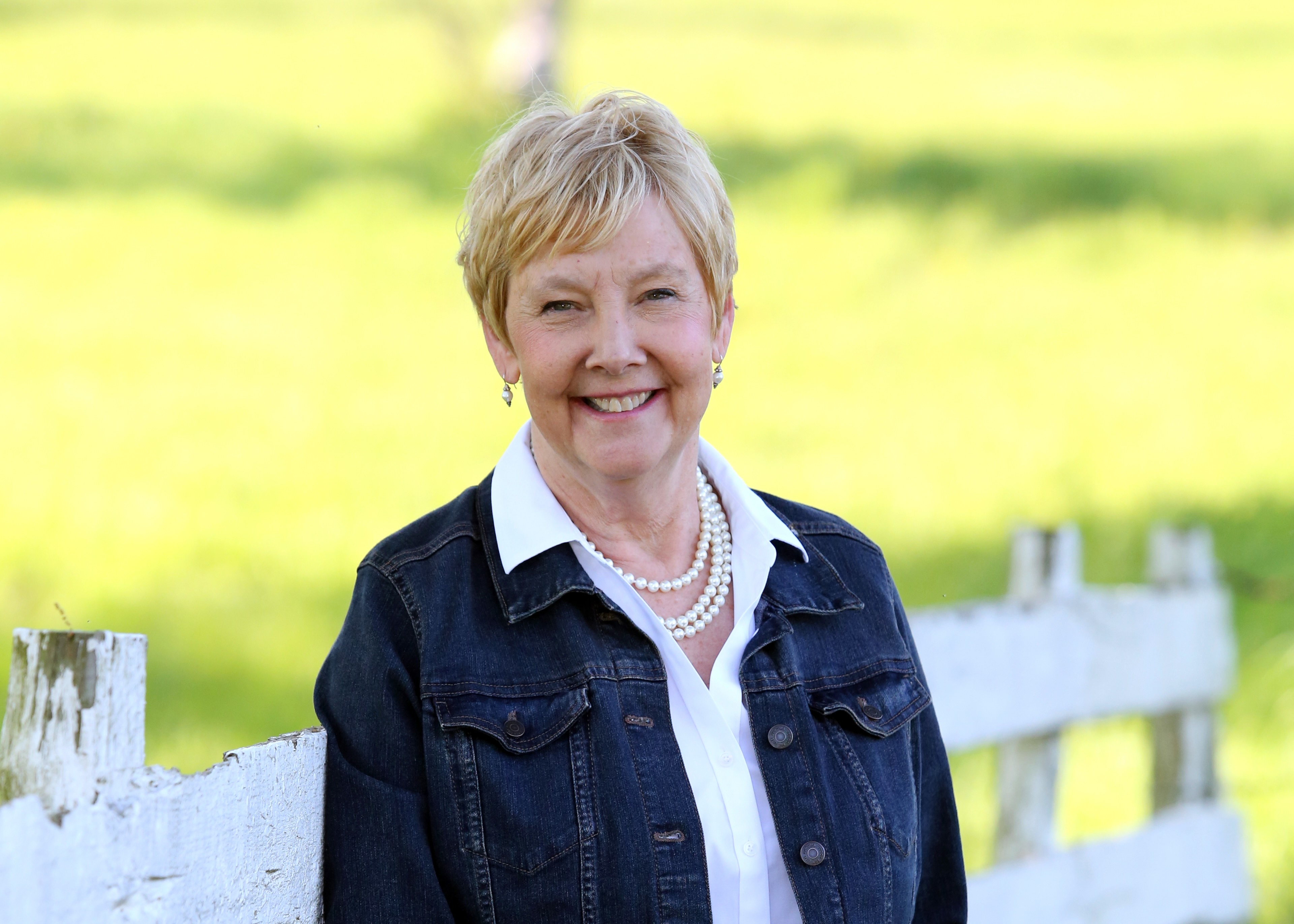 woman with short hair, blue sweater and pearls smiling, standing in a field with a fence