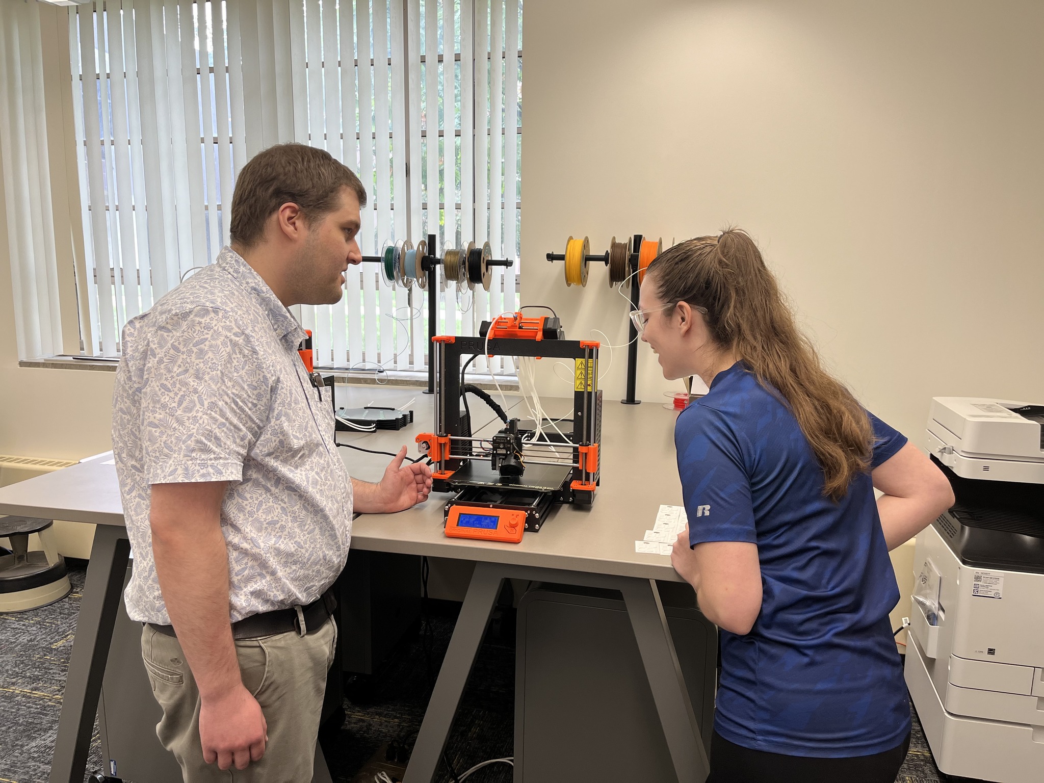 Man with short dark hair watches a 3 D printer. A student stands to his right.