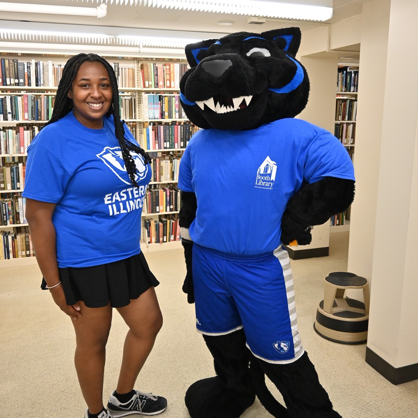 A smiling student with long dark braids stands next to the Billy Panther mascot. Billy has a large black panther head with sharp teeth and a blue highlight from their ears to their cheeks. Billy is wearing a blue tee shirt with a whit Booth Library logo. They are wearing blue shorts with a white stripe. The student is wearing a blue E I U tee shirt with a Panther logo and a black skirt.