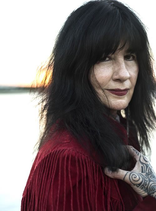 Joy Harjo wearing dark red lipstick and a dark red shirt. There are designs on her hand.