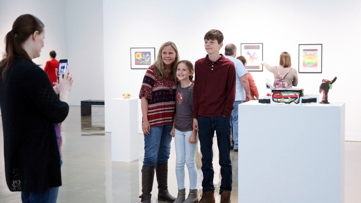 3 people smile as another woman takes a picture of them next to their artwork