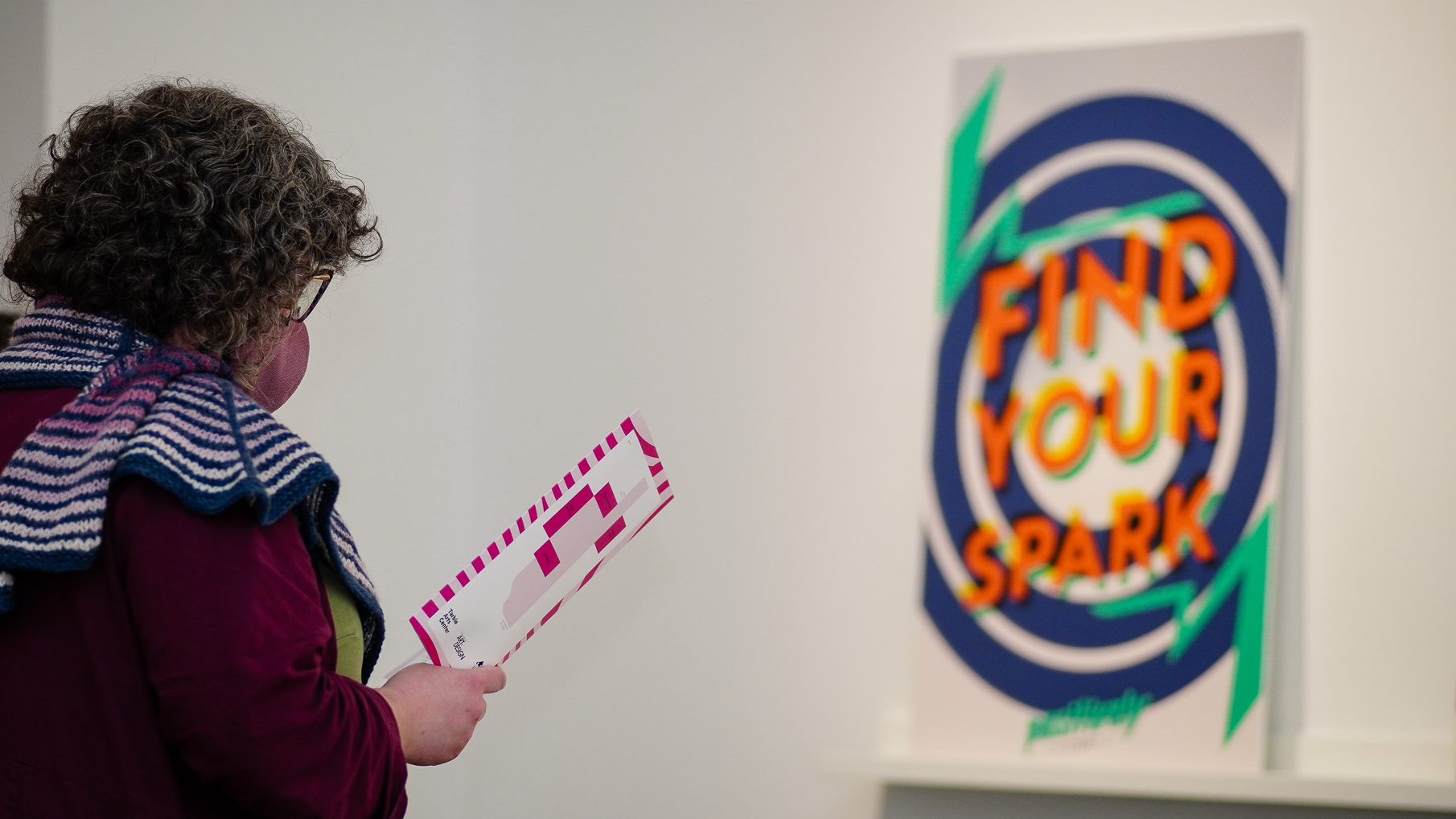 A woman stands holding a gallery guide looking at a large poster that says "Find Your Spark"